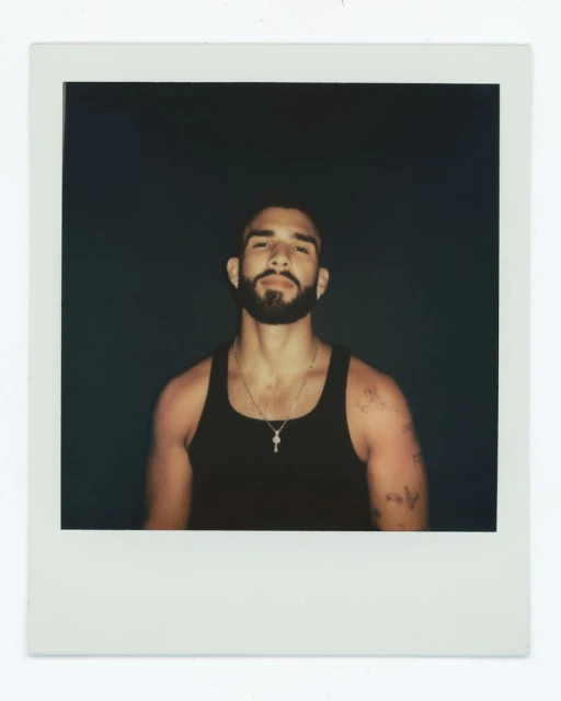 Polaroid portrait of an adult male with a beard wearing a tank top against a dark background, showing shoulders and chest.