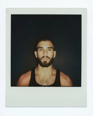 Polaroid portrait of musician Manuel Turizo wearing a black tank top and silver chain necklace against a dark background.