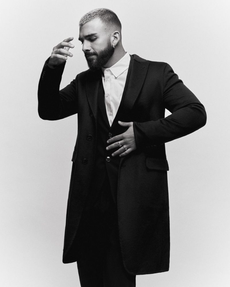 Portrait of a stylish adult male with a beard and short, dyed hair, wearing a black coat, vest, and white shirt, against a solid backdrop.