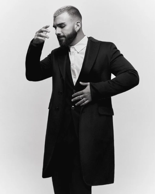 Portrait of a stylish adult male with a beard and short, dyed hair, wearing a black coat, vest, and white shirt, against a solid backdrop.