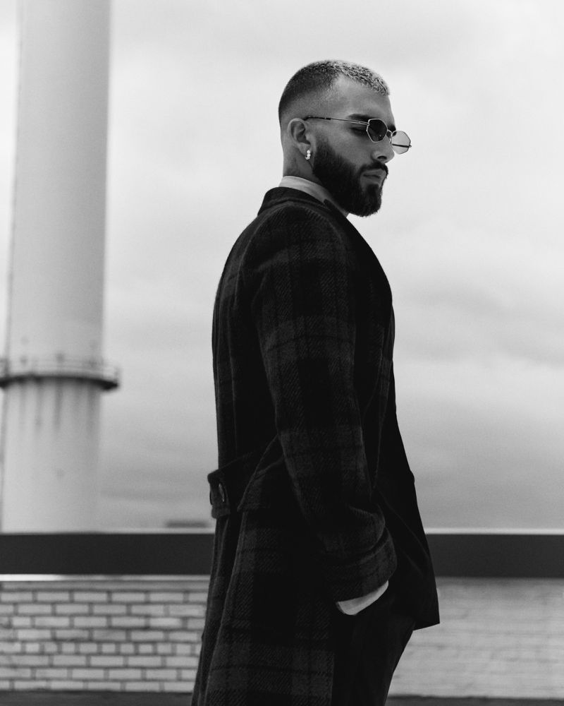Monochrome portrait of a stylish man with a beard wearing sunglasses and a coat, standing on a rooftop with a cloudy sky.