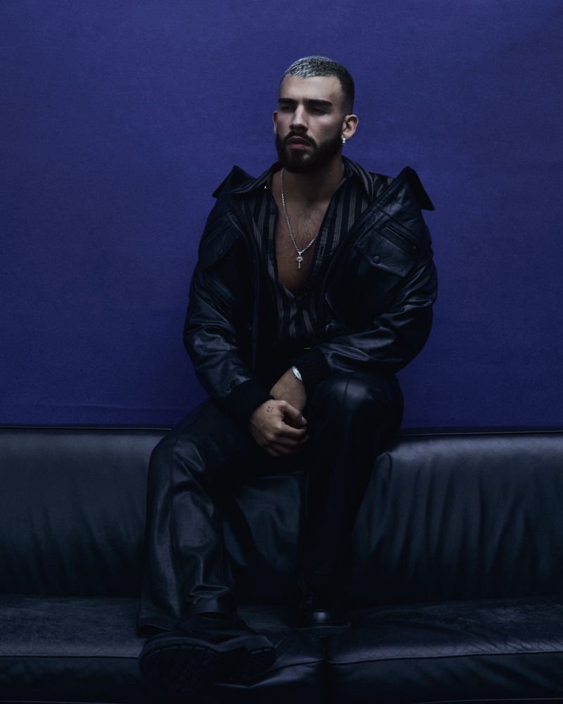Stylish man with salt-and-pepper buzz cut sits on a black couch against a dark purple background, wearing a black leather jacket and pants.