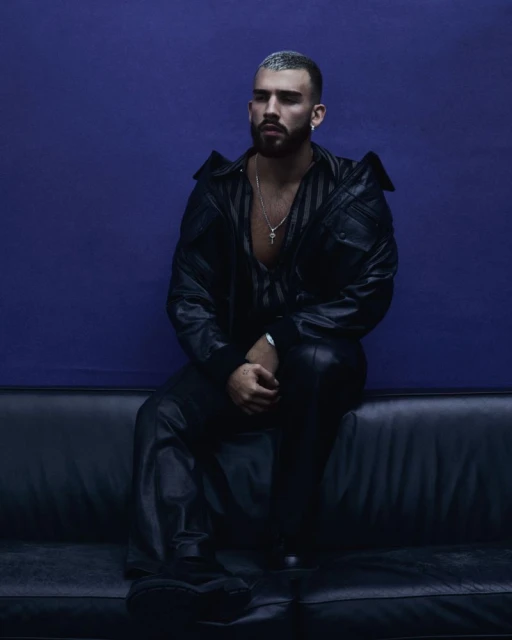 Stylish man with salt-and-pepper buzz cut sits on a black couch against a dark purple background, wearing a black leather jacket and pants.