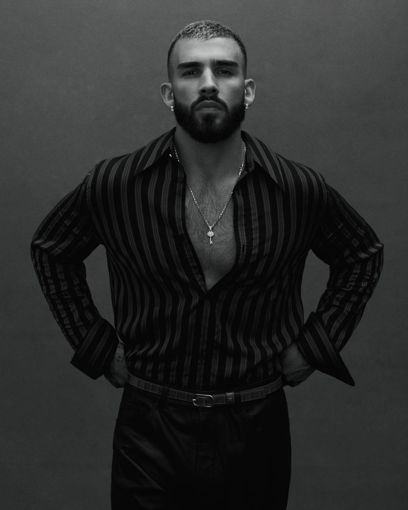Black and white portrait of a confident man with a beard in a striped shirt and black pants posing indoors against a gray background.