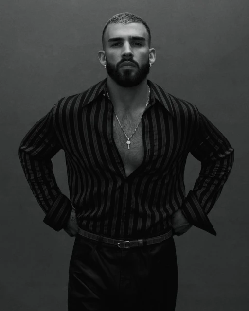 Black and white portrait of a confident man with a beard in a striped shirt and black pants posing indoors against a gray background.