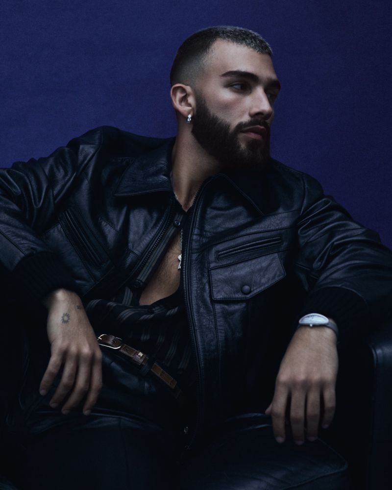 Stylish adult male with beard and short hair poses in black leather jacket, black pants, and silver jewelry against a blue background.