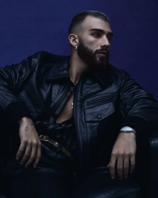 Stylish adult male with beard and short hair poses in black leather jacket, black pants, and silver jewelry against a blue background.