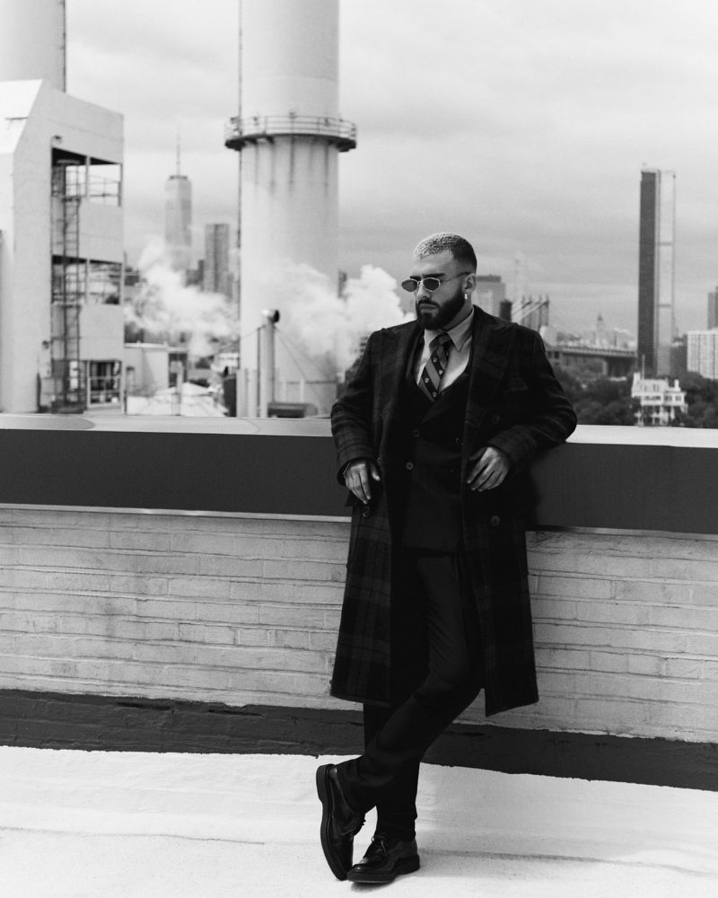 Monochrome portrait of an adult man in a suit and overcoat standing on a rooftop with a cityscape in the background.