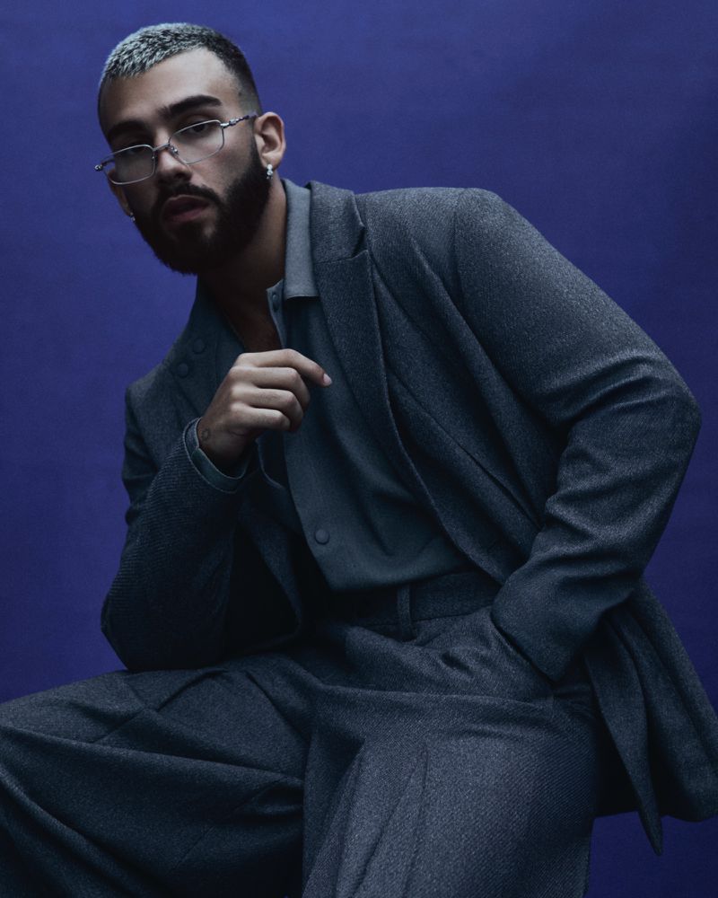 Portrait of a stylish adult male in a gray suit and eyeglasses, seated against a dark blue background in a studio shot.