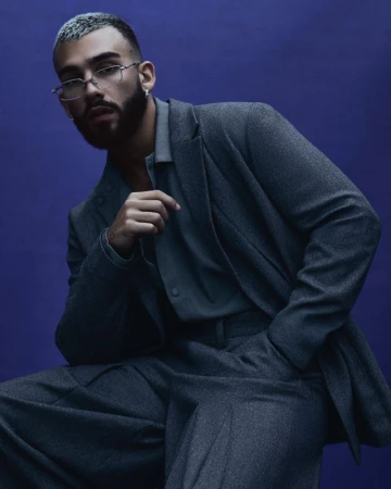 Portrait of a stylish adult male in a gray suit and eyeglasses, seated against a dark blue background in a studio shot.