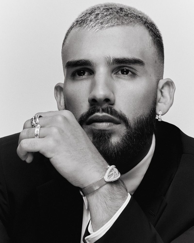 Portrait of an adult male with short bleached hair, a beard, wearing a black suit, and jewelry posing in a photo studio with a light backdrop.