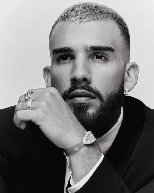 Portrait of an adult male with short bleached hair, a beard, wearing a black suit, and jewelry posing in a photo studio with a light backdrop.