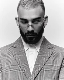 Man wearing round glasses, a plaid suit jacket, and white shirt against a white seamless background