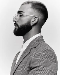 Profile portrait of Manuel Turizo wearing glasses and a plaid suit against a white background for Numero Netherlands.