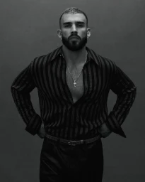 Portrait of a man wearing a dark striped button-down shirt and silver chain necklace against a textured dark background.