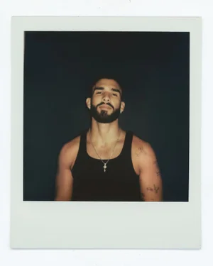 Polaroid photograph of Manuel Turizo wearing a black tank top and a silver key pendant necklace against a dark background.