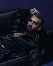 Man with beard and short hair wearing a black leather jacket resting on a dark sofa in a moody studio setting