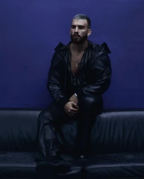 Man with beard seated on a dark leather couch wearing a black leather jacket against a blue background