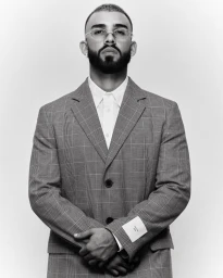 Man wearing a plaid suit jacket and white shirt posing against a plain grey background for Numero Netherlands.
