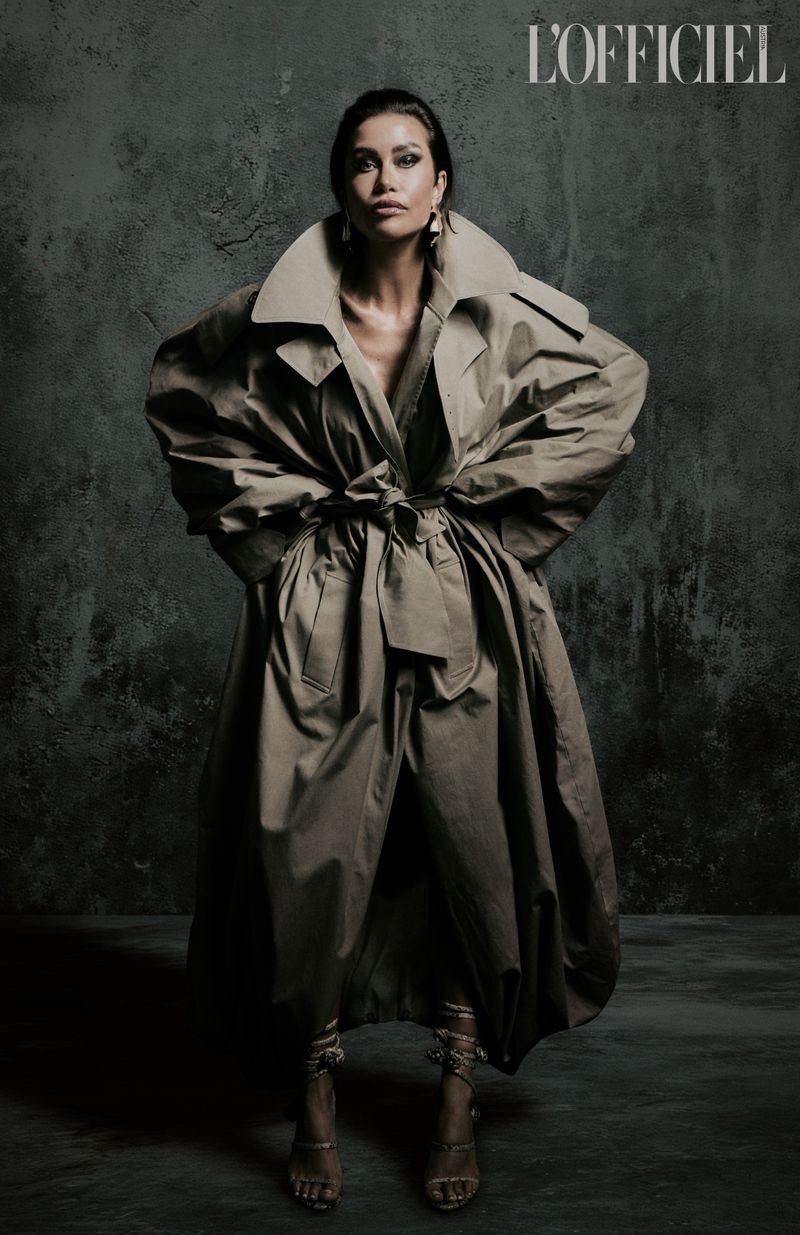 A fashion portrait features a woman in a voluminous trench coat against a textured grey backdrop, with the L'Officiel logo visible.