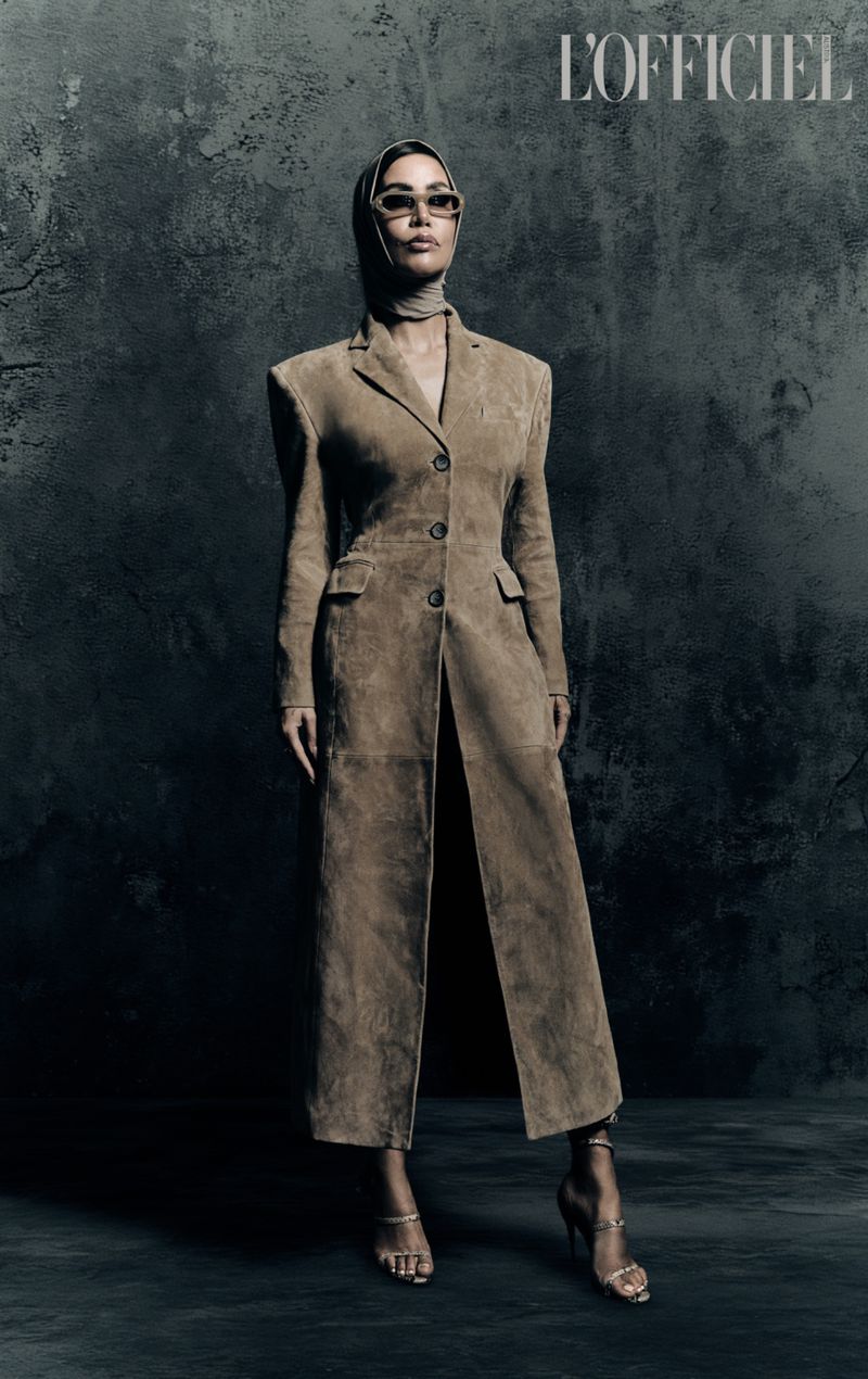 A full-length studio shot features an adult woman in a tan coat and head covering, accessorized with jewelry and heels, set against a dark backdrop.