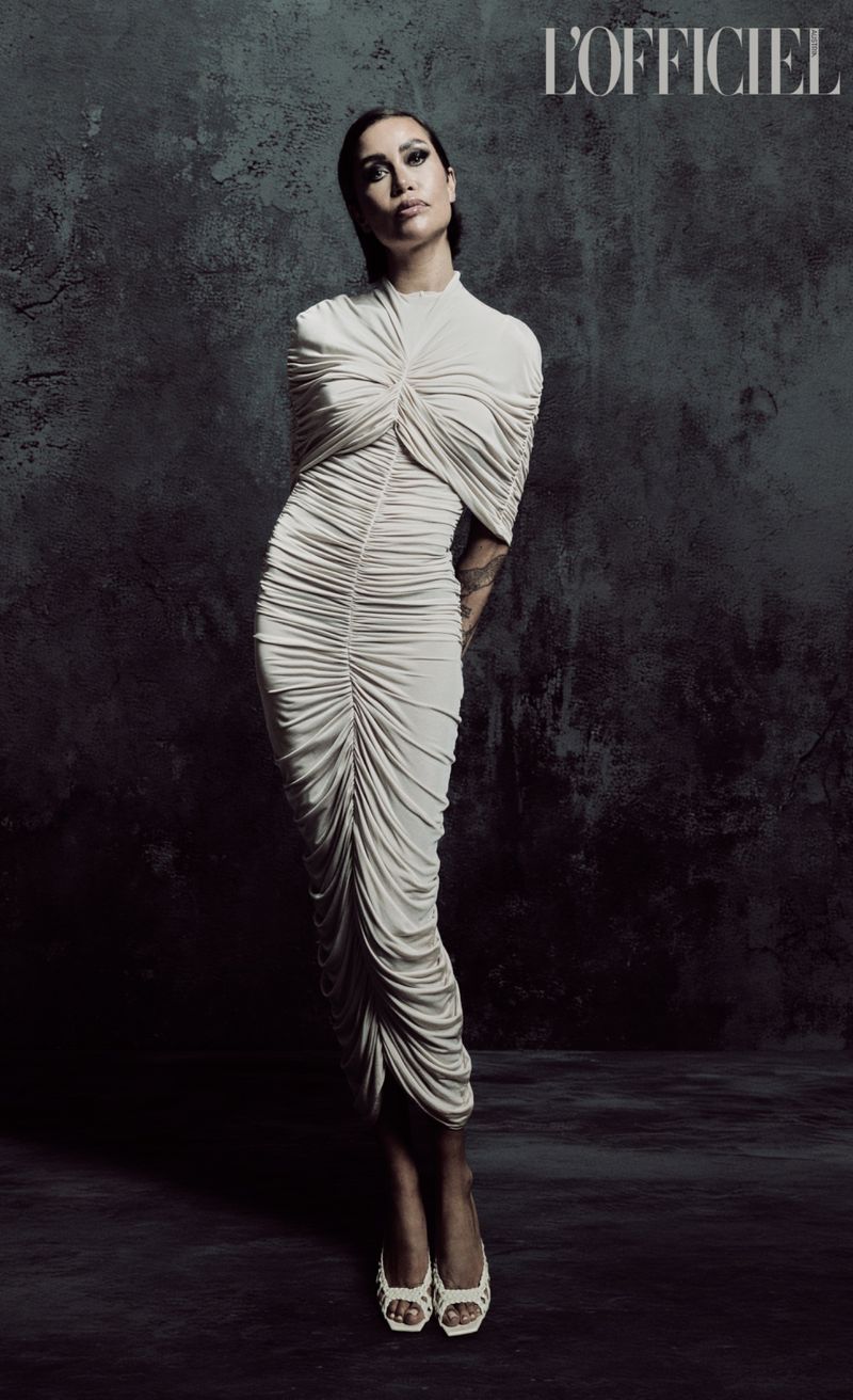 Full-length shot of a woman in a white draped dress and matching heels against a dark grey mottled backdrop. She poses looking forward.