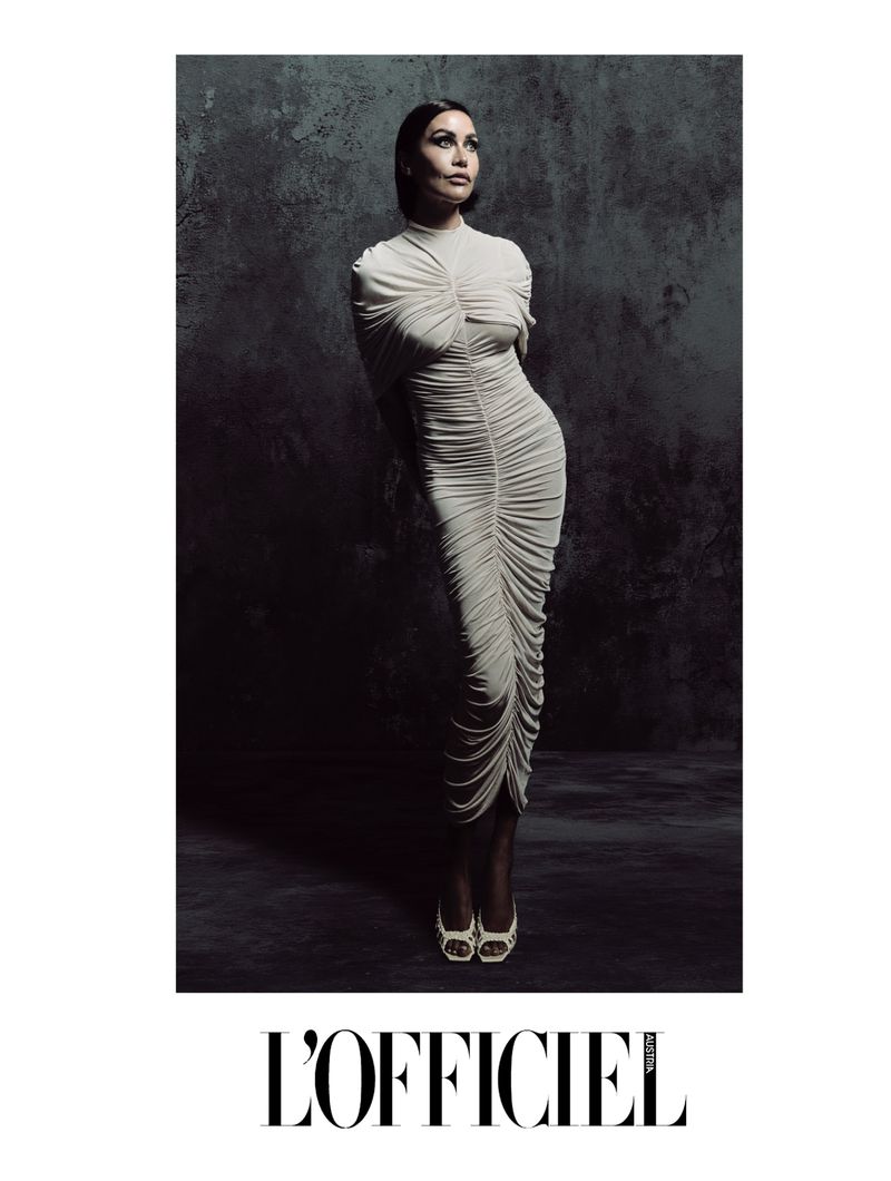 A woman in a light dress poses against a textured background, featured in L'Officiel magazine. The image uses soft, directional lighting.
