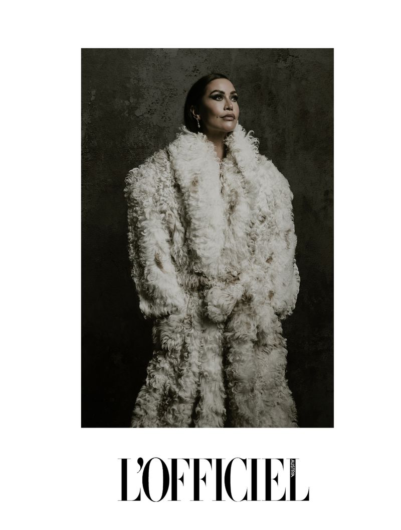 Portrait of a woman in a full-length, fluffy white coat, gazing upward against a mottled dark background. L'Officiel logo at bottom.