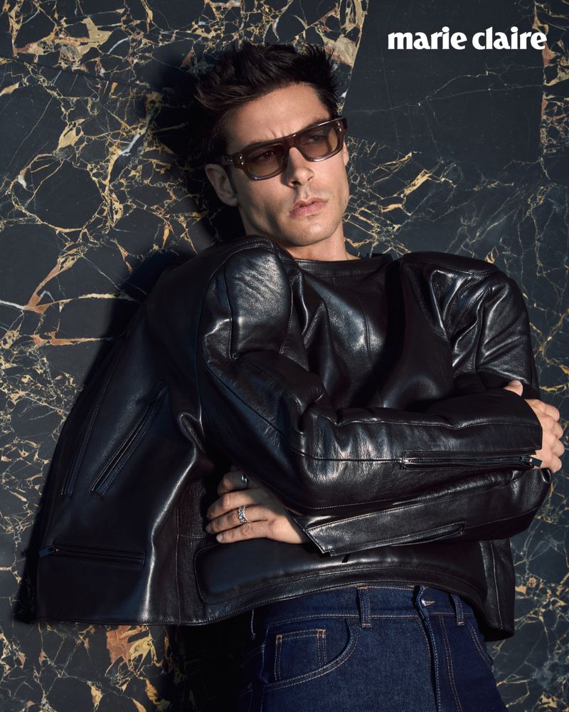 Man in sunglasses and leather jacket poses against a marble backdrop for Marie Claire magazine, showcasing fashion and editorial style.