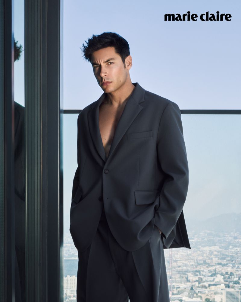 Portrait of a brunette adult male wearing a dark suit with an open collar, posed in front of a window with a city view.