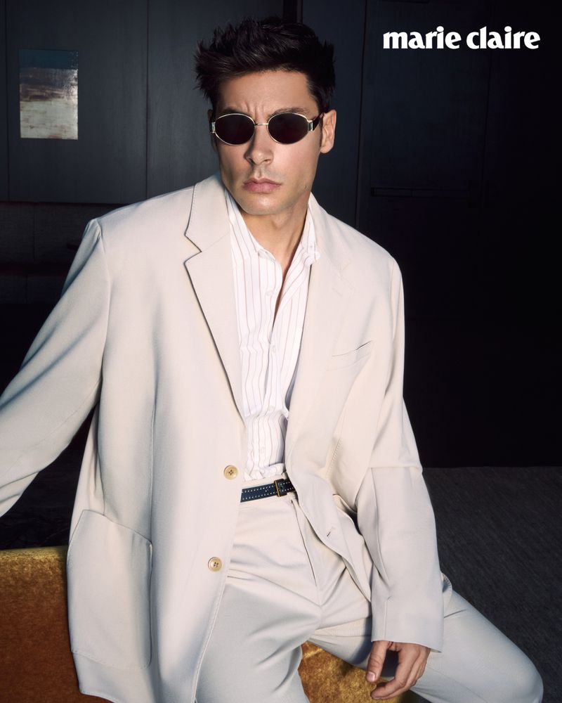 Portrait of a stylish man in sunglasses, an off-white suit, and an unbuttoned striped shirt, posing indoors next to a gold surface.