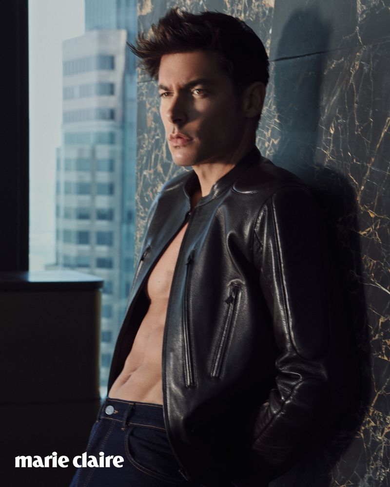 Handsome man poses in leather jacket with building and marble wall background. Fashion editorial style, confident gaze, and masculine style.