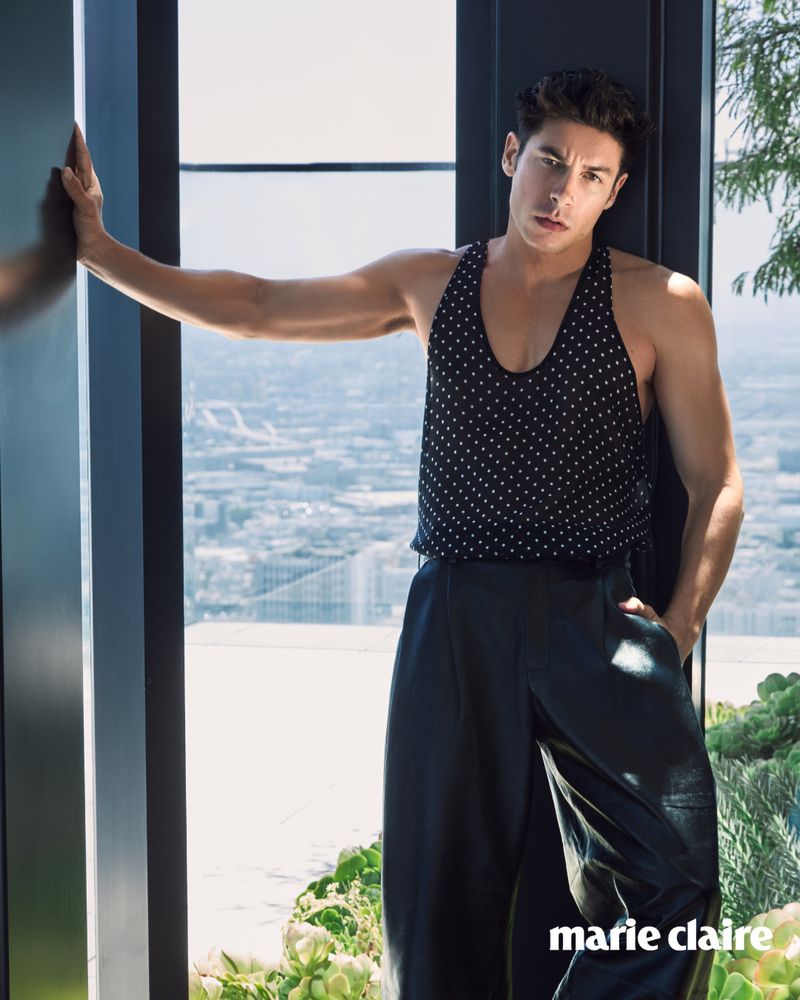 Indoor shot of a slender adult male fashion model posing in a black polka-dot tank top and baggy pants near a window with city views.