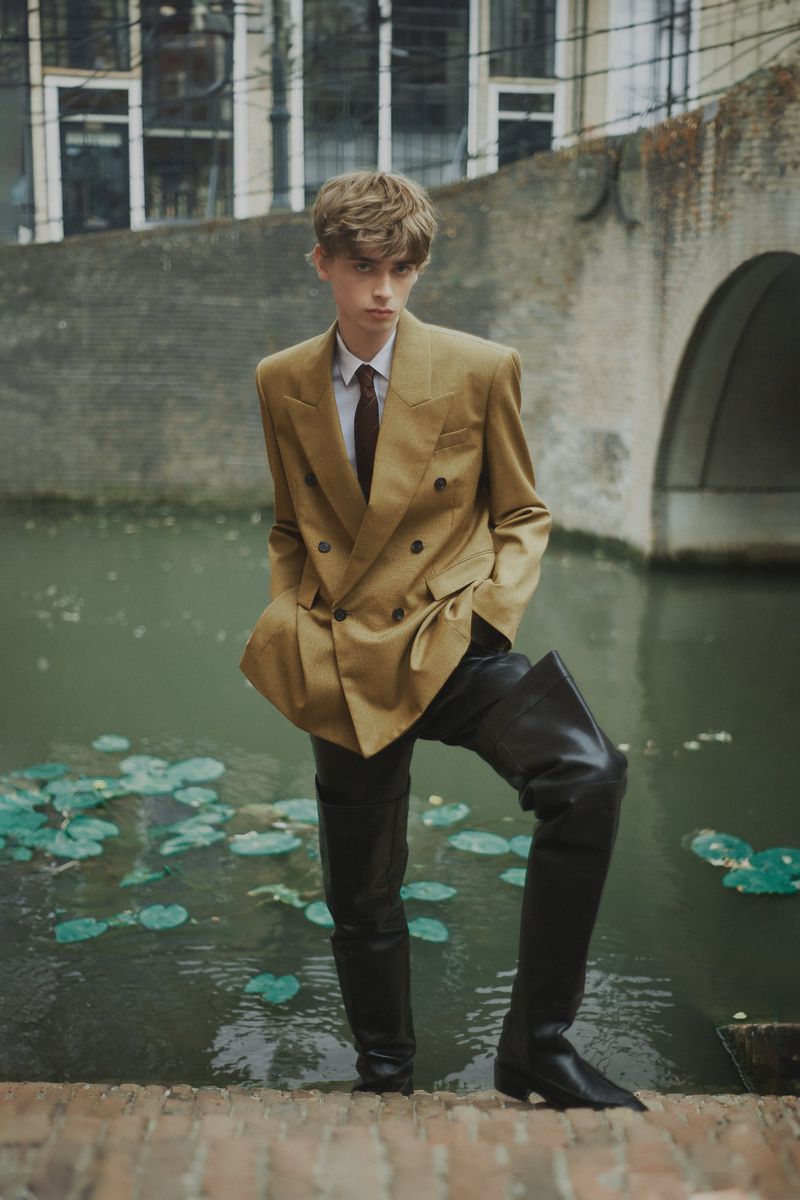 A young adult in a tan double-breasted blazer and black leather boots poses on stone steps near a canal with water lilies.