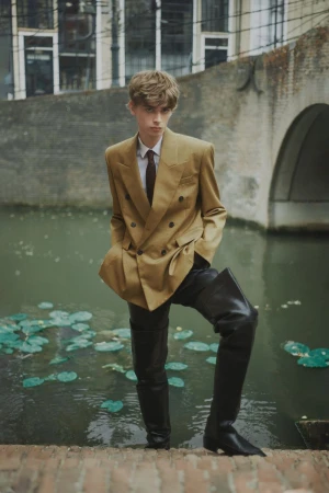 A young adult in a tan double-breasted blazer and black leather boots poses on stone steps near a canal with water lilies.