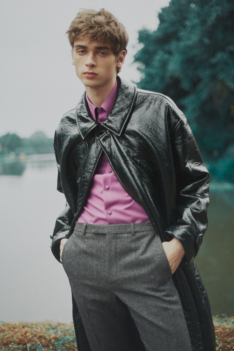 Male fashion model poses outdoors wearing a black patent leather coat, pink shirt, and gray trousers, with a lake and trees background.