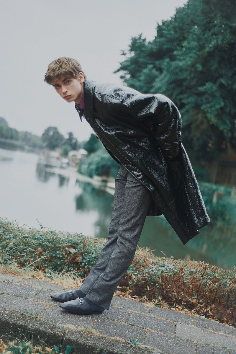 A young adult male model with brown hair poses outdoors near water, wearing a long black coat, gray pants, and black shoes, showcasing fashion.