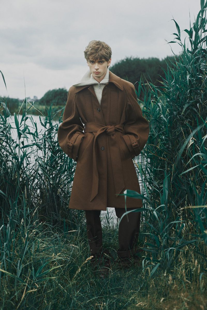 A young adult poses in a brown trench coat by a lake surrounded by tall reeds against an overcast sky in a fashion editorial.