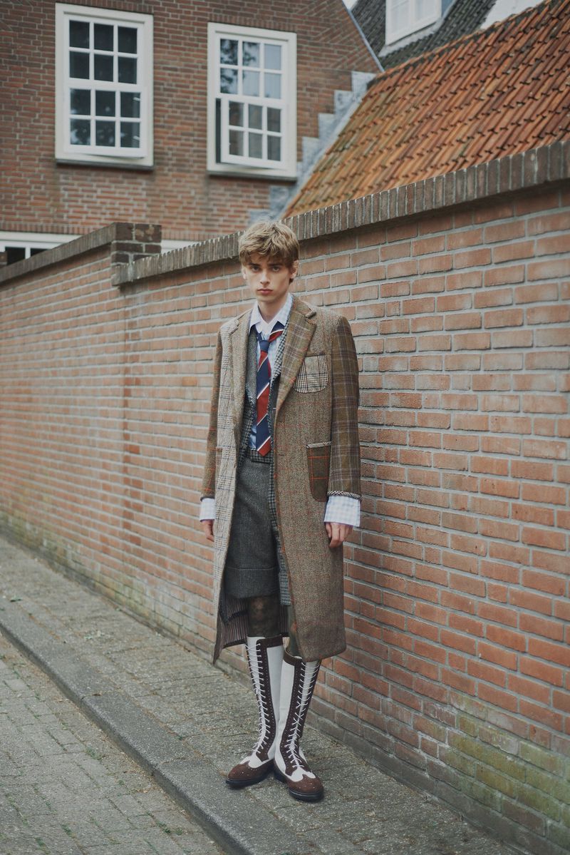 Male fashion model in coat, tie, shorts, and high socks poses against a brick wall in an outdoor, urban setting with natural lighting.
