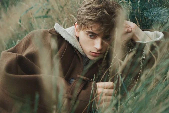Portrait of a young man lying in tall grass. He wears a brown jacket with a gray hoodie, gazing off with a subtle expression.