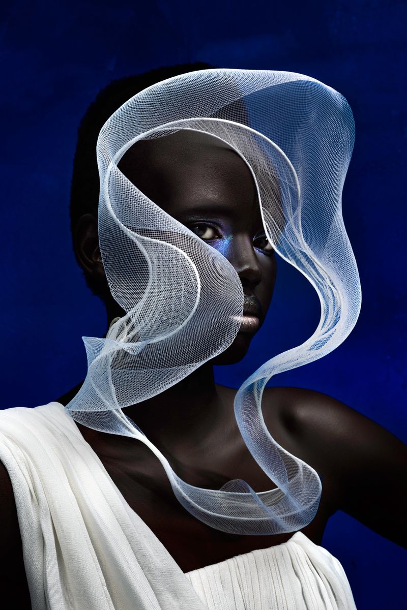 Portrait of an African woman with blue eye makeup, wearing a white draped dress and a sculptural white net veil against a blue backdrop.
