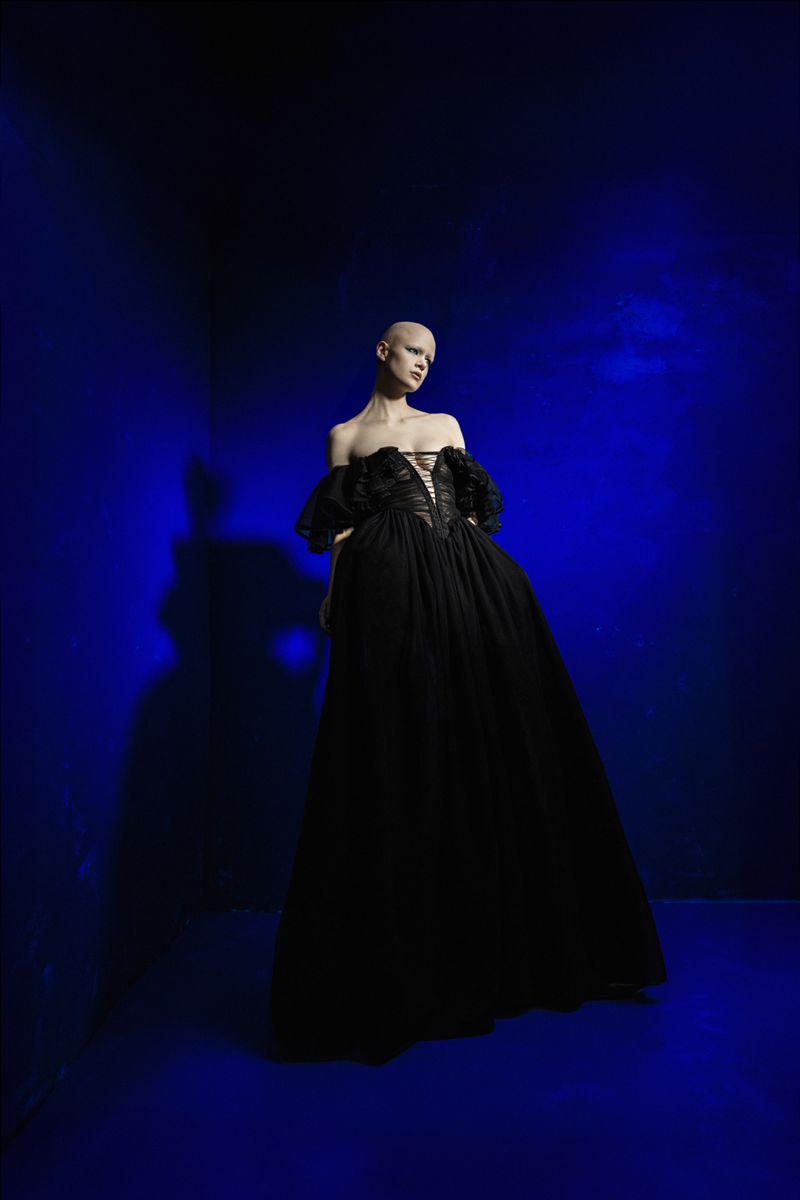A person wearing a black gown poses against a striking blue backdrop, captured in a fashion-style full shot with studio lighting.