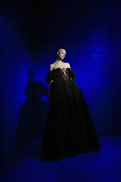 A person wearing a black gown poses against a striking blue backdrop, captured in a fashion-style full shot with studio lighting.