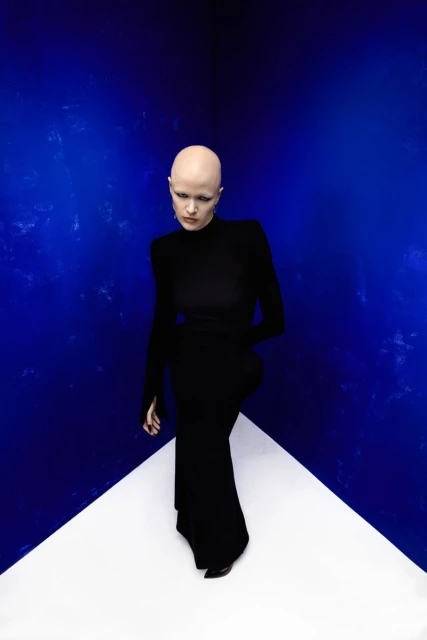 An androgynous, bald adult stands centered in a long black dress against a geometric blue and white studio backdrop in portrait orientation.