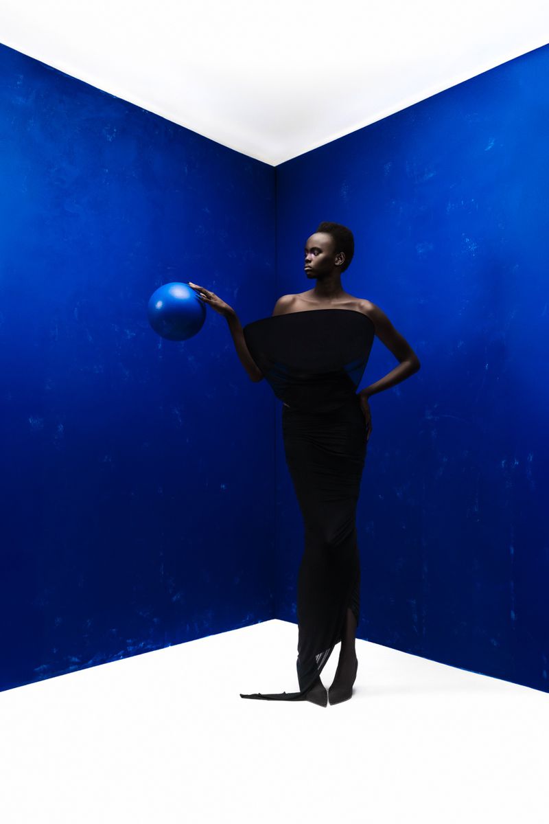 Elegant African woman in black dress holding a blue sphere in a photo studio with a blue wall and white floor.