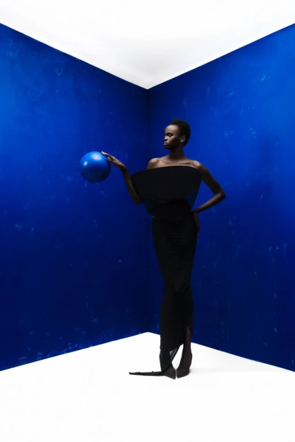 Elegant African woman in black dress holding a blue sphere in a photo studio with a blue wall and white floor.