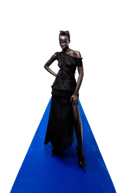 A slender fashion model with dark skin and blue eye makeup poses confidently on a blue surface against a white backdrop in a studio shot.
