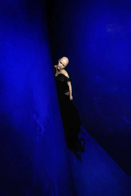 A bald woman in a black dress stands in a dark space illuminated with blue light, creating a striking and artistic fashion portrait.