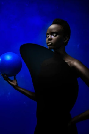 Artistic portrait of a black woman with slicked-back hair holding a blue sphere, complemented by a dark, curved object and blue background.