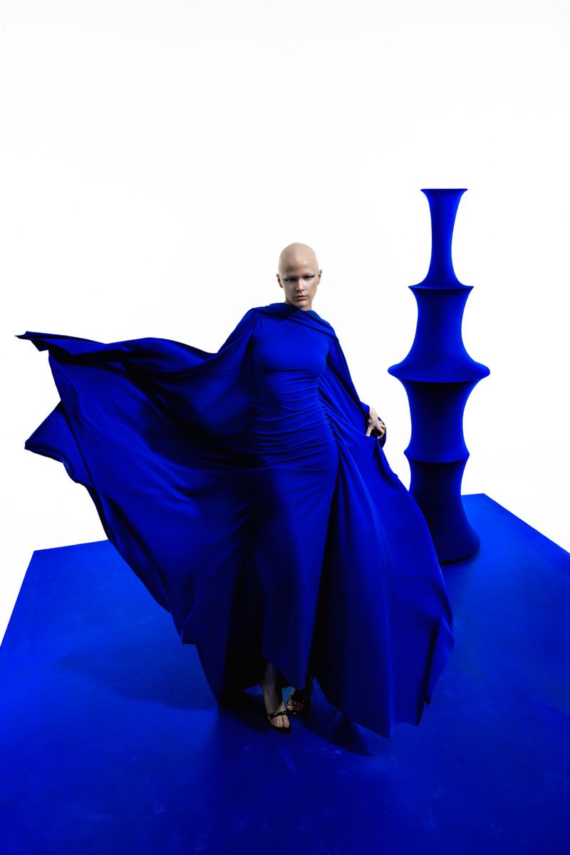 Woman in blue gown posing on a blue platform next to a blue geometric vase in a brightly lit photo studio. Elegant fashion.